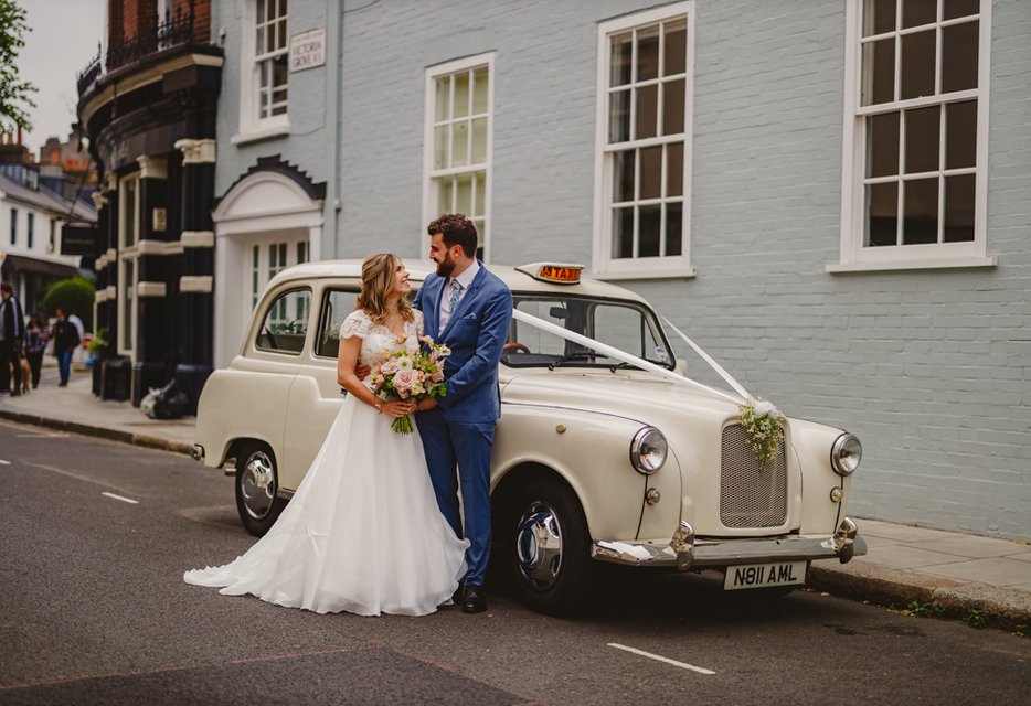 A bride and groom stand lovingly beside a vintage white taxi adorned with flowers on a london city street. The scene exudes romance and elegance from a chelsea wedding photographer.