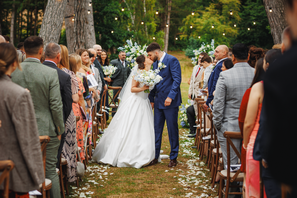 Bride and groom kissing outdoors under string lights at brookfarm wedding venue, surrounded by guests on either side of an aisle adorned with white flower petals, conveying joy and celebration.
