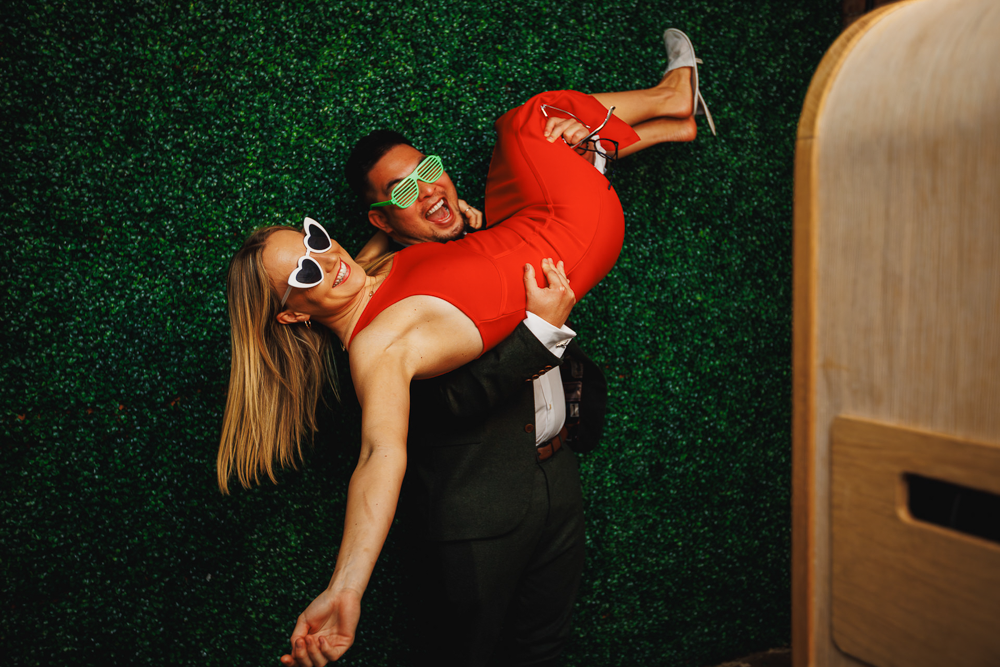 A man in a suit playfully lifts a woman in a red dress against a green backdrop. Both wear fun sunglasses and are smiling, conveying joy and celebration.