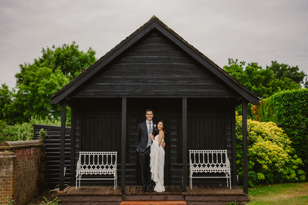 A couple stands joyfully in front of a rustic black wooden cabin on the grounds of micklefield hall wedding venue, embraced under a cloudy sky. Greenery and white benches frame the cheerful scene.