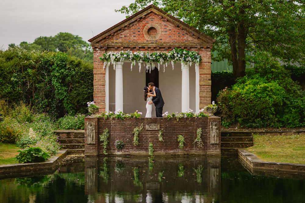 A couple embraces in front of a small, brick neoclassical building adorned with white columns and floral garlands, reflected in a calm pond. Romantic atmosphere at micklefield hall wedding venue.