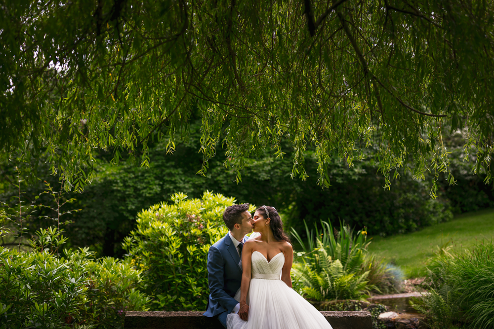 A couple shares a romantic kiss under a lush, green canopy the garden of pennyhill park wedding venue. The bride wears a white gown, and the groom a blue suit, surrounded by vibrant foliage.