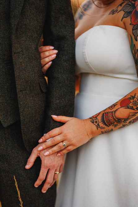 A bride in a white dress with colorful tattoos gently holds the arm of a groom in a dark suit. Both wear rings, conveying love and commitment.