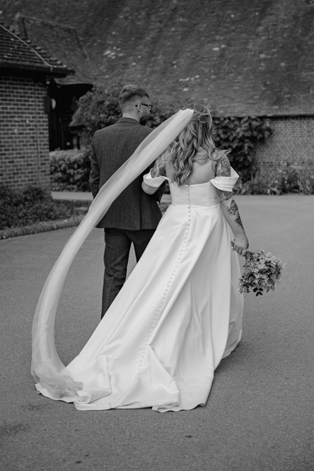 A bride in a flowing gown and veil walks hand-in-hand with a groom in a suit, both facing away. The scene is peaceful and romantic.