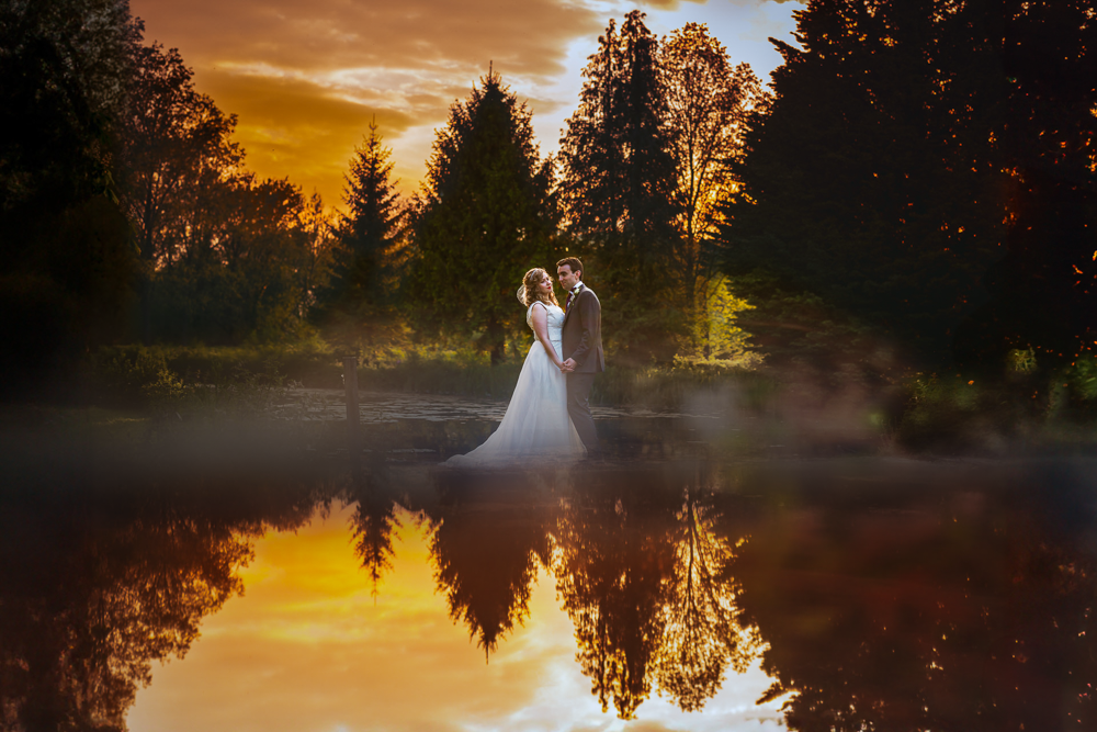 A couple in wedding attire embrace by a reflective lake at sunset at the old mill wedding venue. The sky is vibrant with orange and purple hues, and silhouetted trees add a serene backdrop.