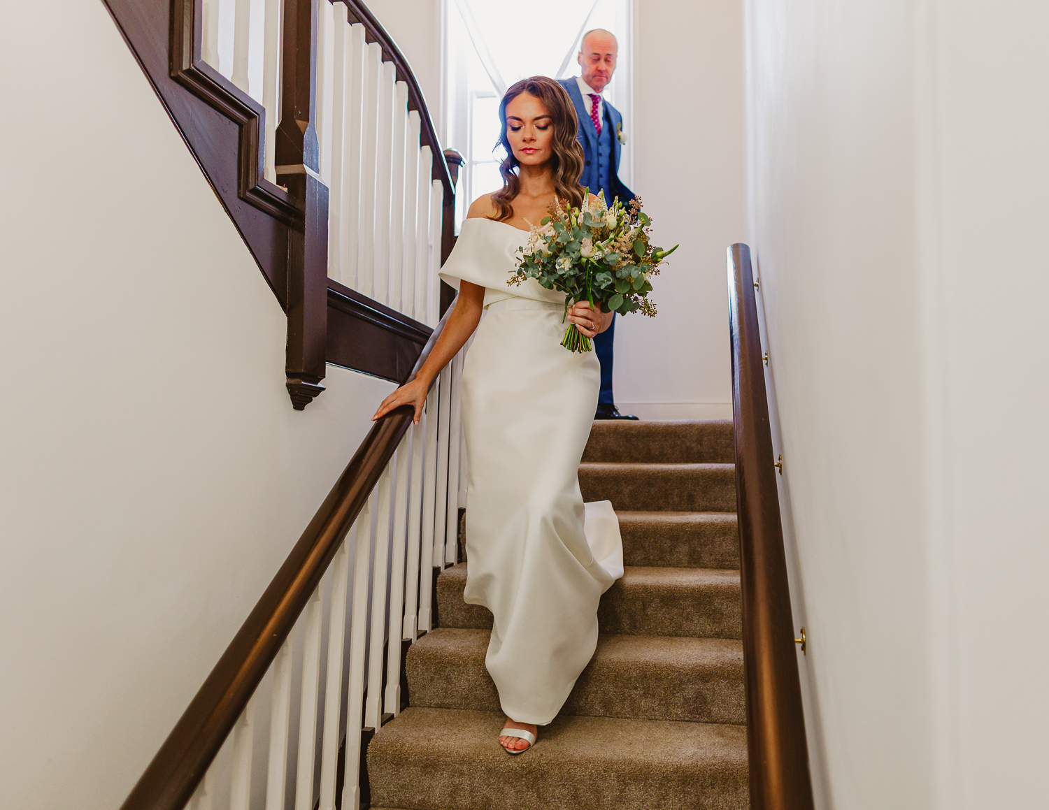 A bride in an elegant white dress descends a staircase, holding a bouquet of greenery. A man follows behind. The scene is calm and poised.