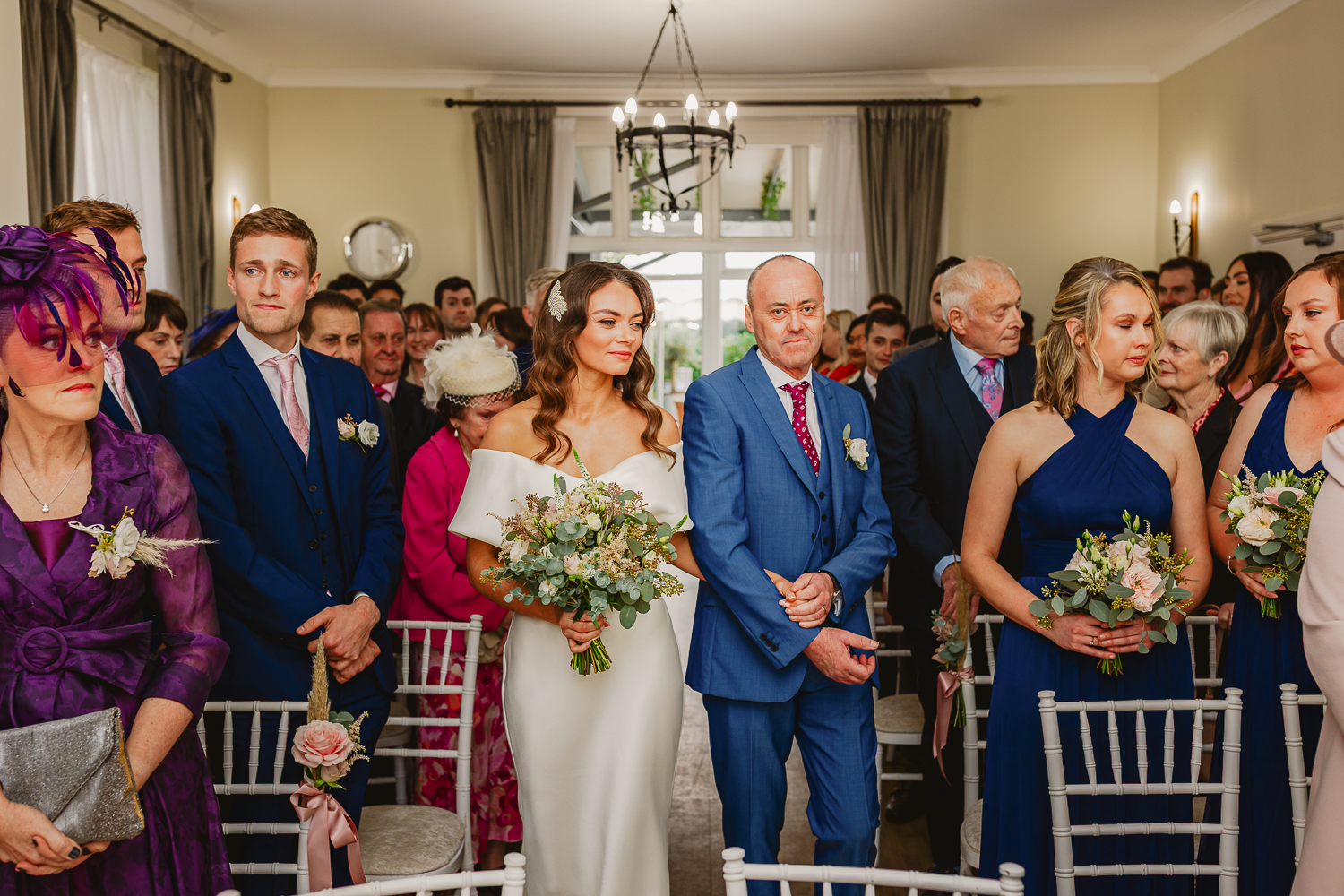 A bride in an elegant white gown and a man in a blue suit stand in a room lined with guests. The scene is festive and formal, with flowers and elegant attire indicating a wedding ceremony.