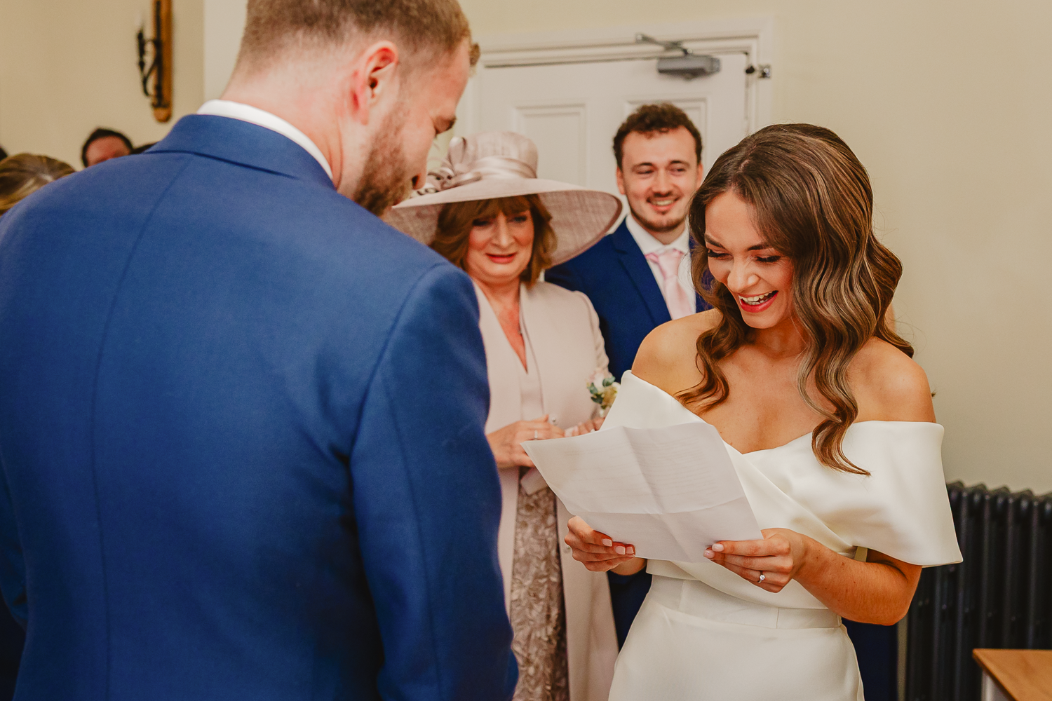 A bride in an off-shoulder white dress joyfully reads a letter to the groom, who is in a blue suit. Smiling guests stand in the background.