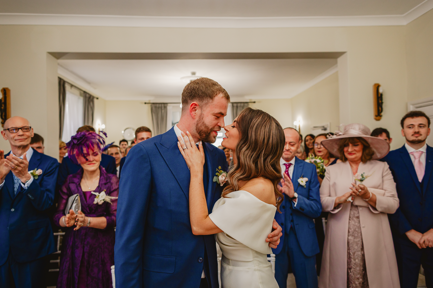 A bride and groom share a joyful kiss indoors, surrounded by applauding guests. The room is warmly lit, and the atmosphere is celebratory and intimate.