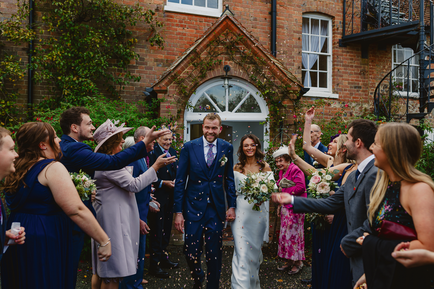 A bride and groom in formal attire walk joyfully through a crowd of cheering guests outside a brick building, as confetti is thrown in celebration.
