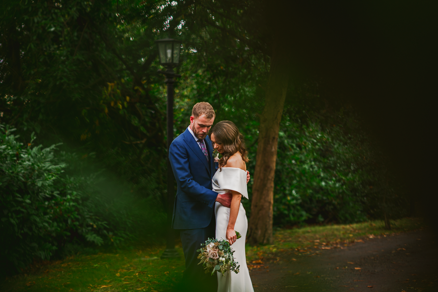 A bride and groom embrace tenderly in a lush garden, surrounded by greenery. The bride holds a bouquet, and both radiate love and serenity.