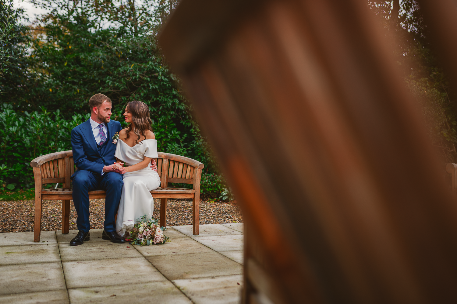 A couple sits on a wooden bench in a garden, holding hands and smiling at each other. The foreground is blurred, adding depth to the intimate scene.