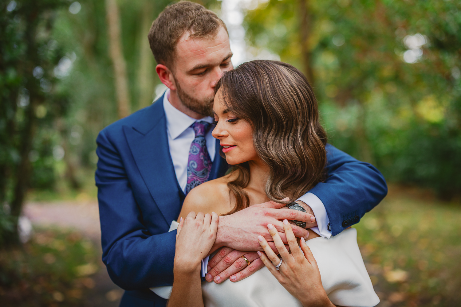 A couple embraces warmly in a lush, green setting. The man in a blue suit gently kisses the woman's head as she smiles, conveying love and serenity.