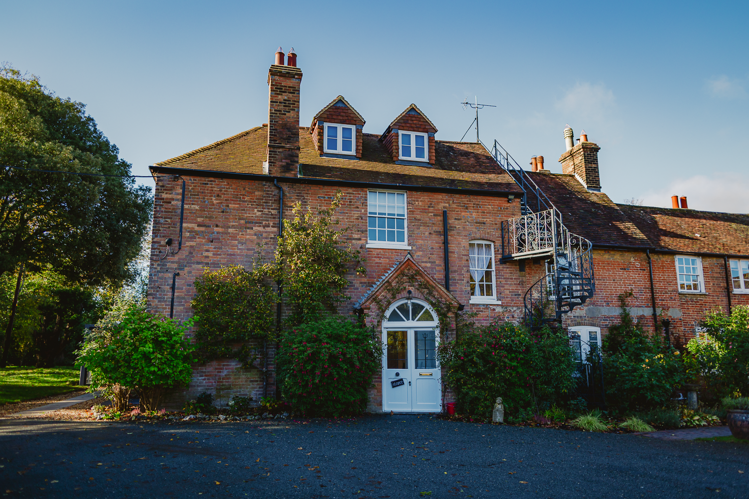The Old Rectory a charming two-story brick house with dormer windows, a white arched door, and a spiral staircase. It's surrounded by lush greenery under a clear blue sky.