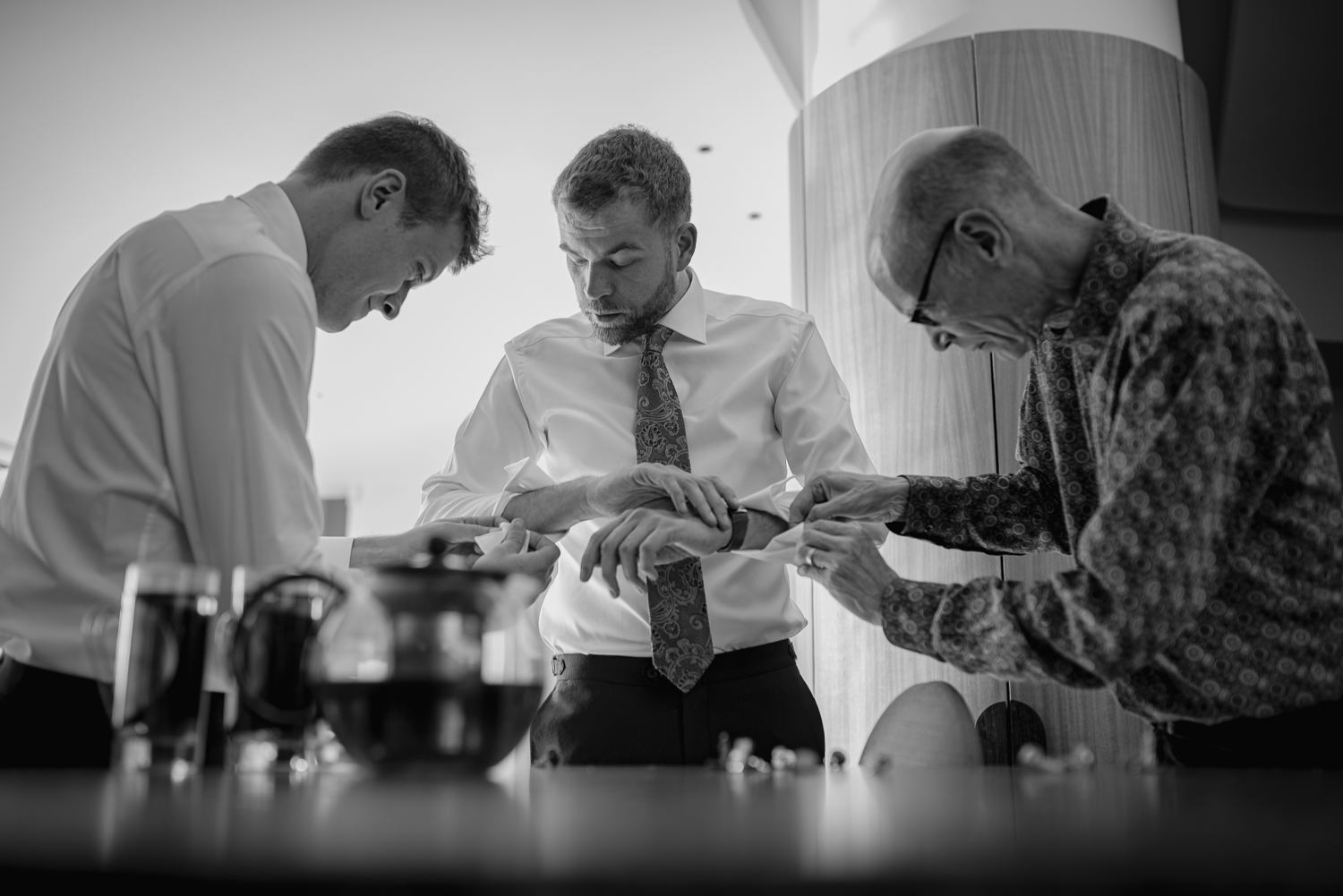 The groom and his best men are interactively putting on watches and cufflinks in a well-lit room. They wear shirts, and focus on their tasks, creating a calm, focused atmosphere.
