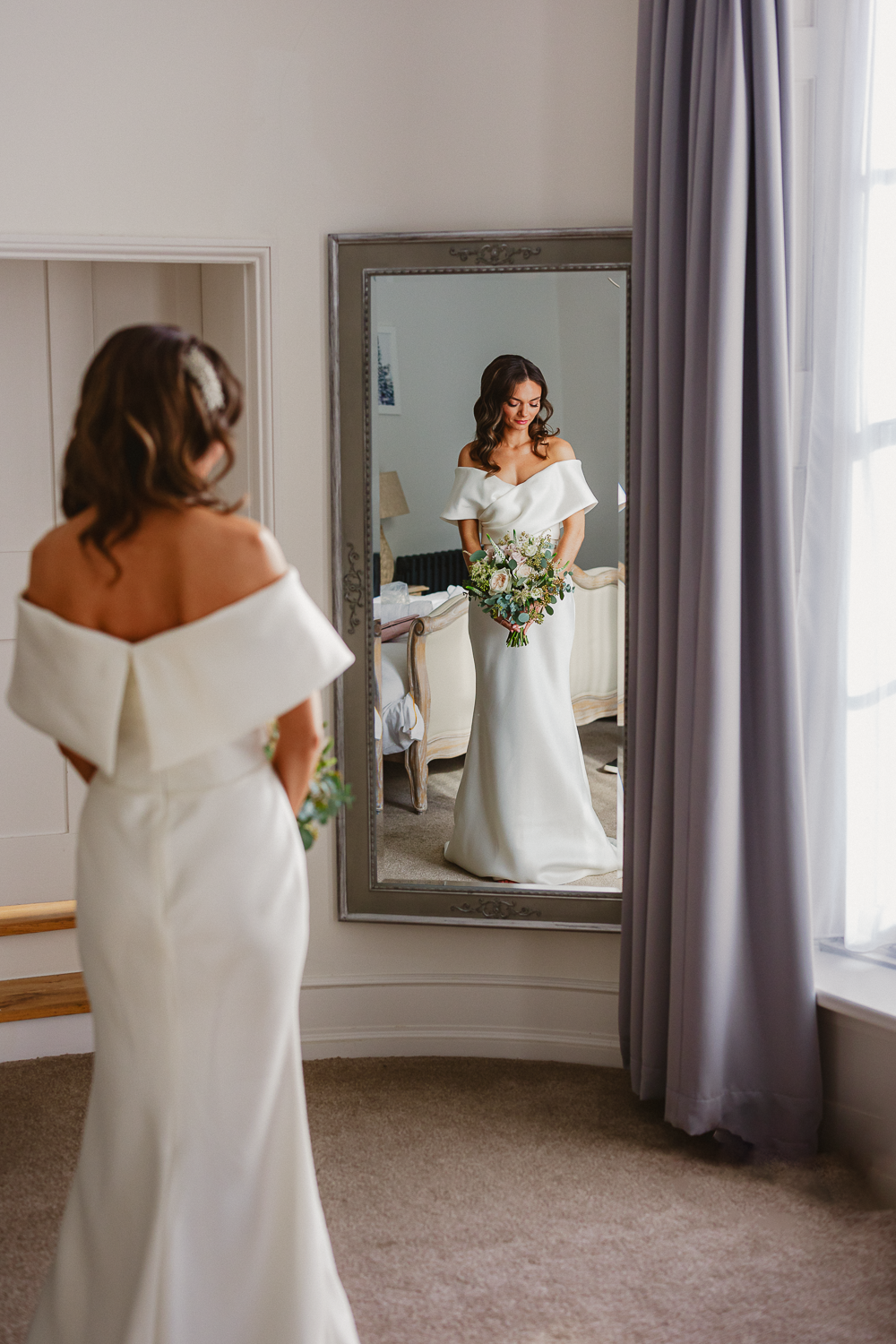 Bride in a white, off-the-shoulder gown holds a bouquet and gazes at her reflection in a mirror, exuding a serene and joyful ambiance.