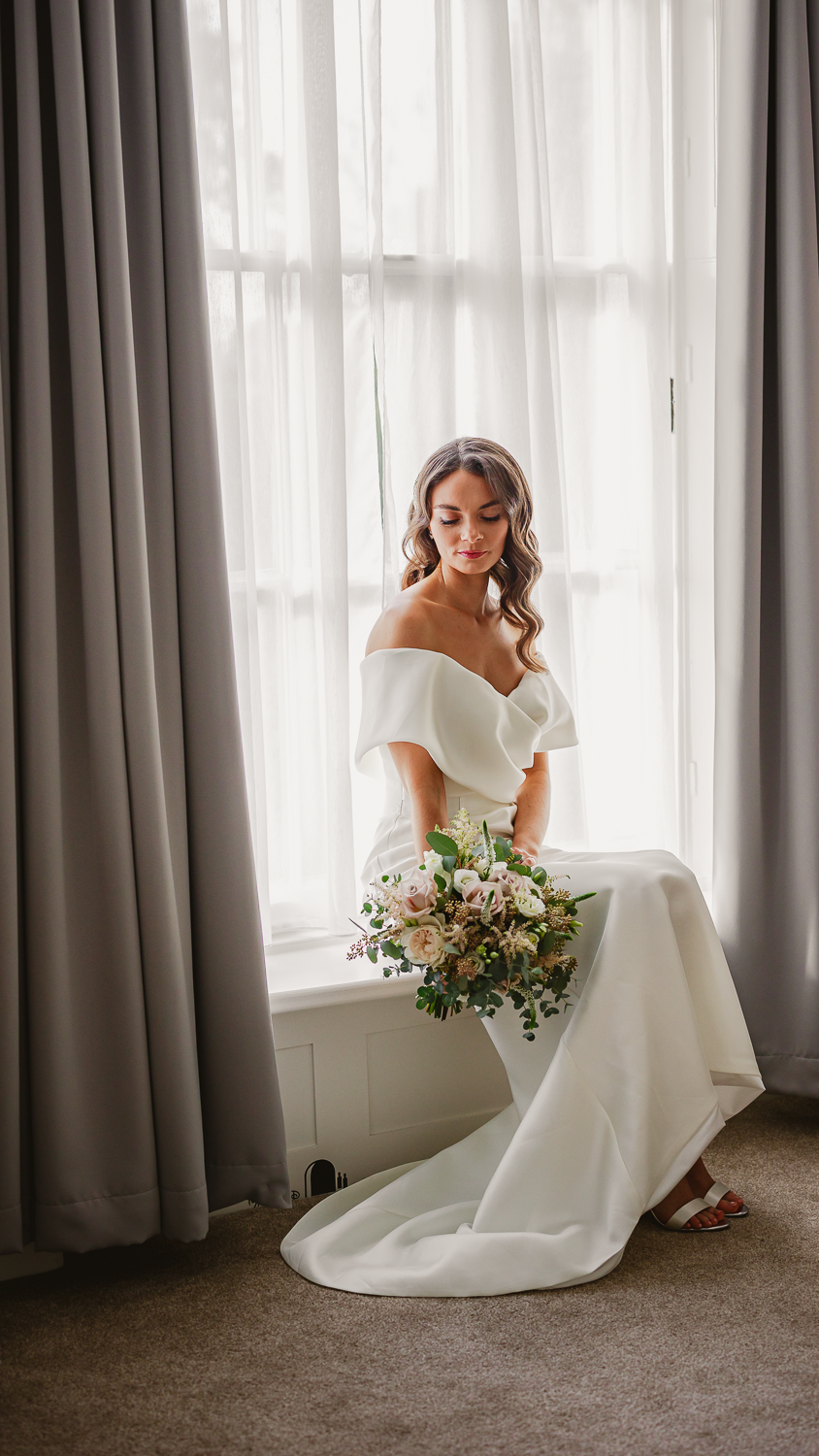 A bride in an off-shoulder white gown holds a bouquet, seated by a window with sheer and gray curtains. She has long hair and a serene expression.