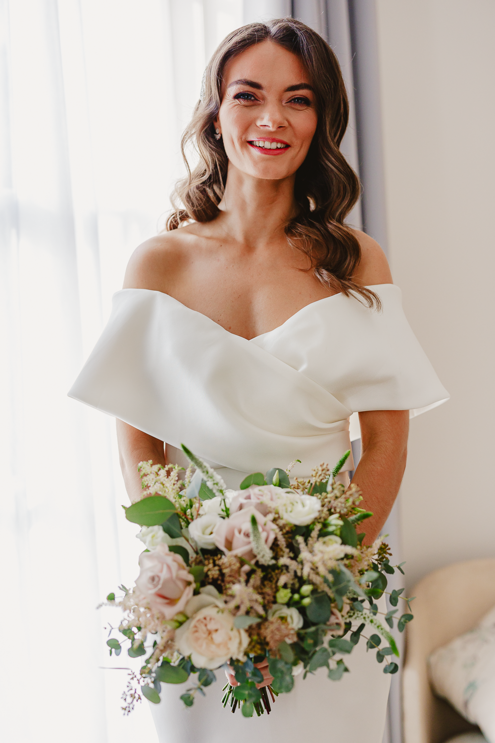 A smiling bride in an elegant off-shoulder white dress holds a lush bouquet with white and pink roses. Soft, natural light adds a warm and joyful ambiance.