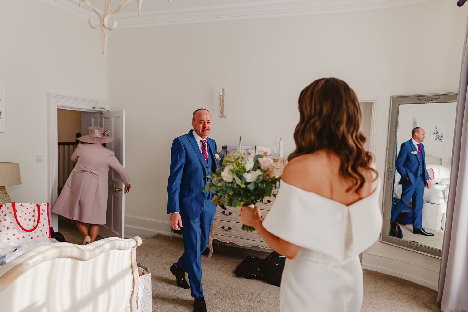 Bride in white dress holds bouquet, facing her dad in a blue suit entering a bright room. A woman in pink exits. Elegant, joyful atmosphere.