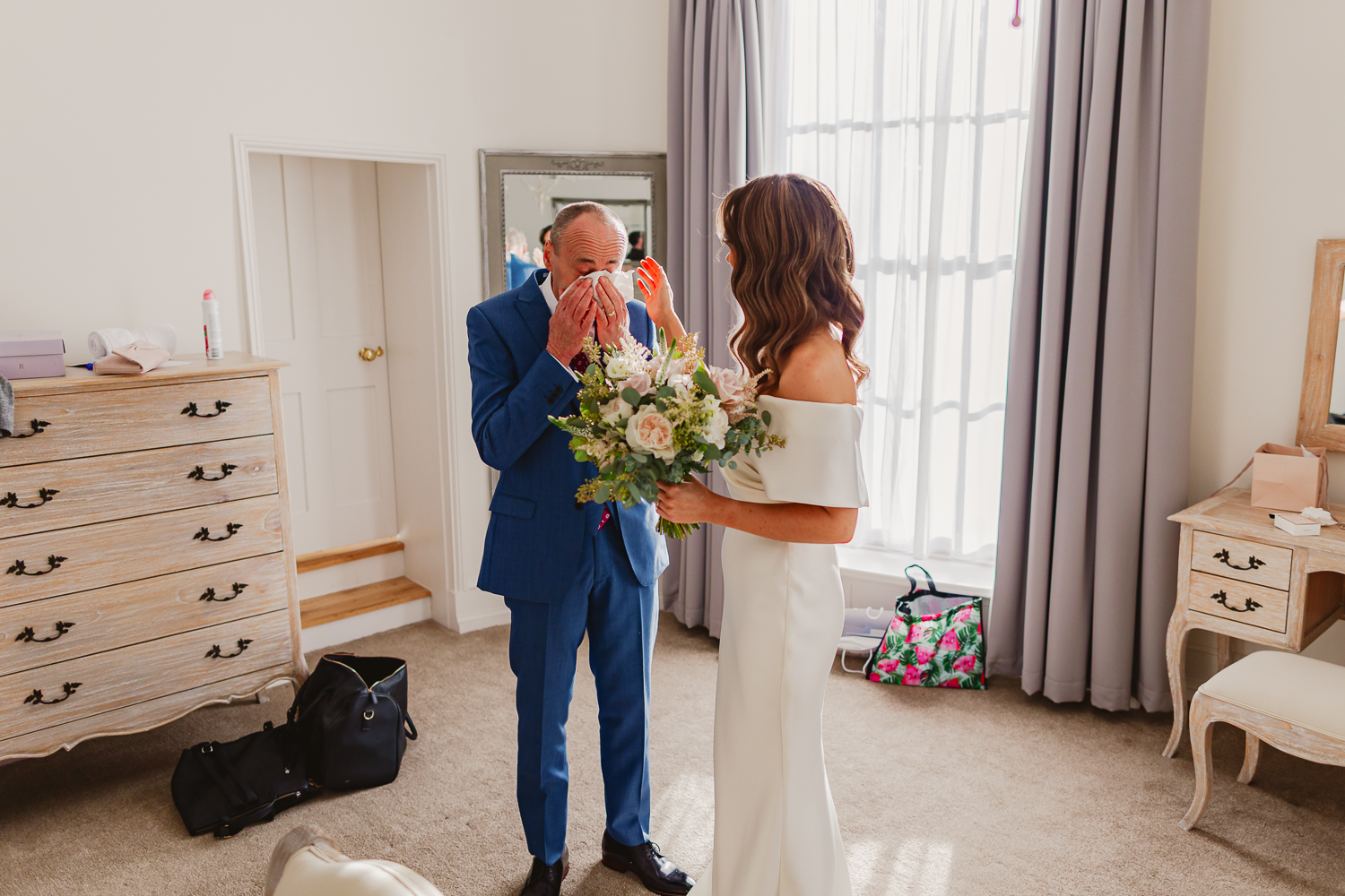 A bride in a white gown holds a bouquet while facing an emotional dad in a blue suit, who wipes tears, in a softly lit room with elegant decor.