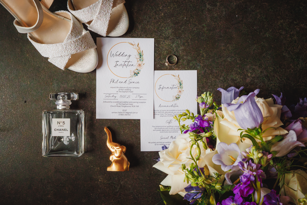 Wedding flat lay with white sandals, floral invitations, a ring, Chanel No. 5 perfume, golden elephant figurine, and a bouquet of purple and cream flowers. Elegant and romantic.