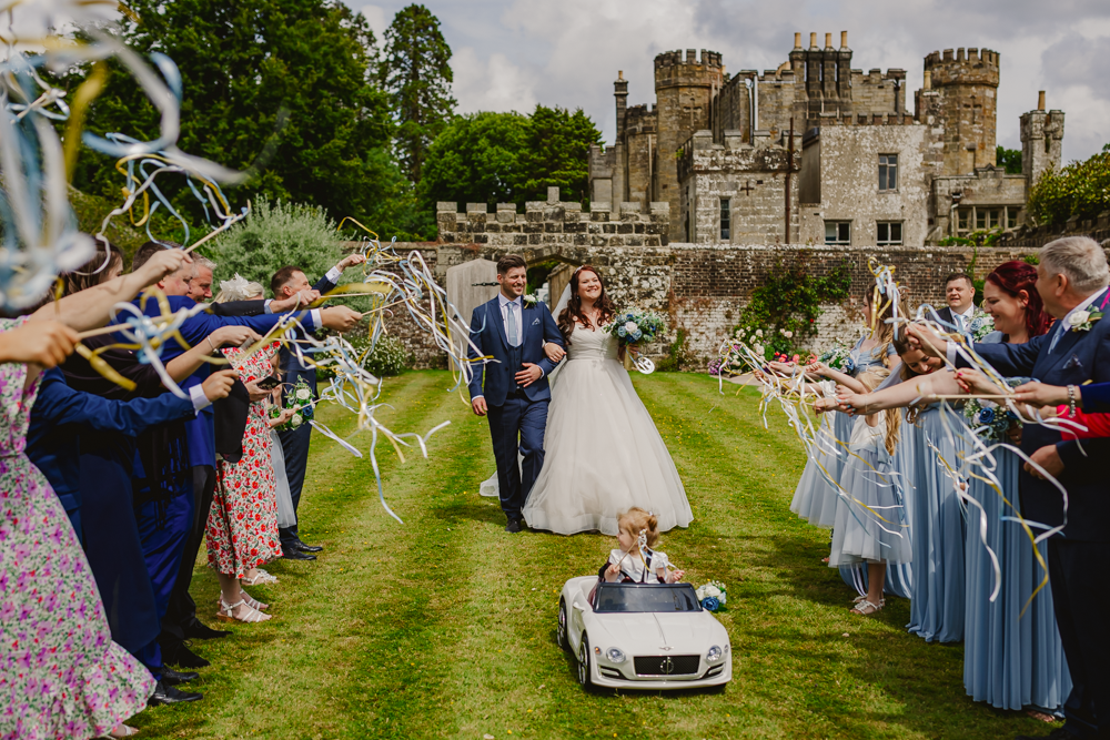 A joyful wedding scene with a couple walking down a grassy aisle outside wadhurst castle, greeted by guests waving ribbons. A child rides a toy car ahead.