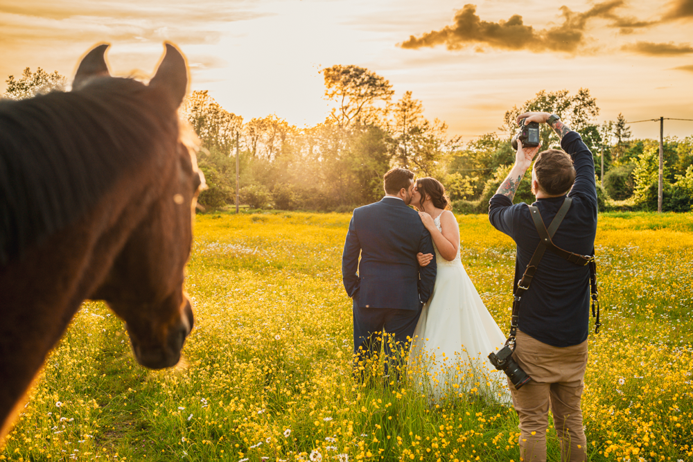 A couple poses in a sunlit field, a photographer captures their embrace, while a horse observes from the foreground, adding a rustic charm.